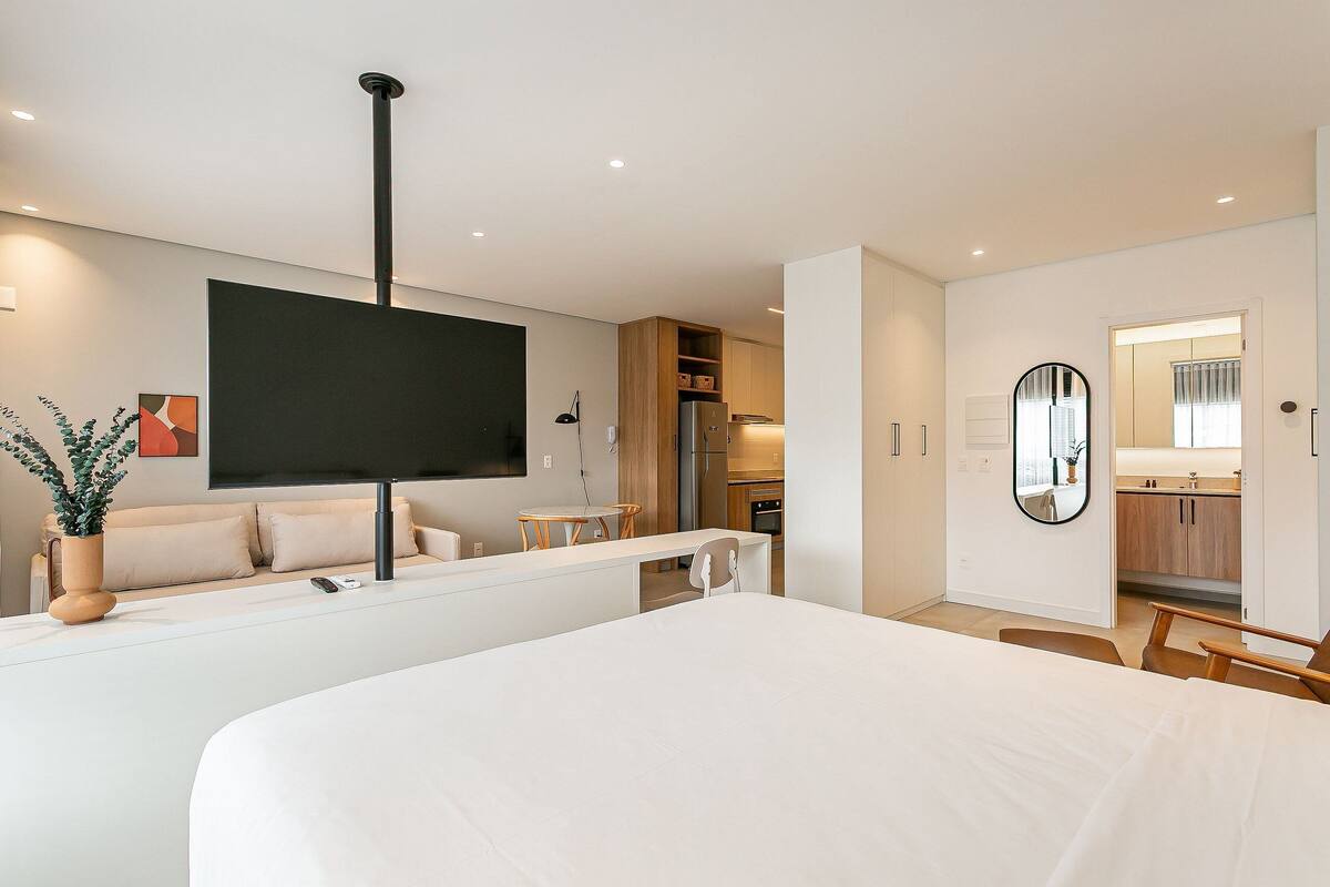 Une chambre d’hôtel moderne avec un grand lit, un téléviseur fixé au mur, un coin repas et une cuisine à l’arrière-plan.