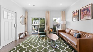 A modern living room with a brown leather sofa, a green armchair, a round coffee table, and a patterned rug. There is a sliding glass door leading to a balcony with a view of buildings and trees.