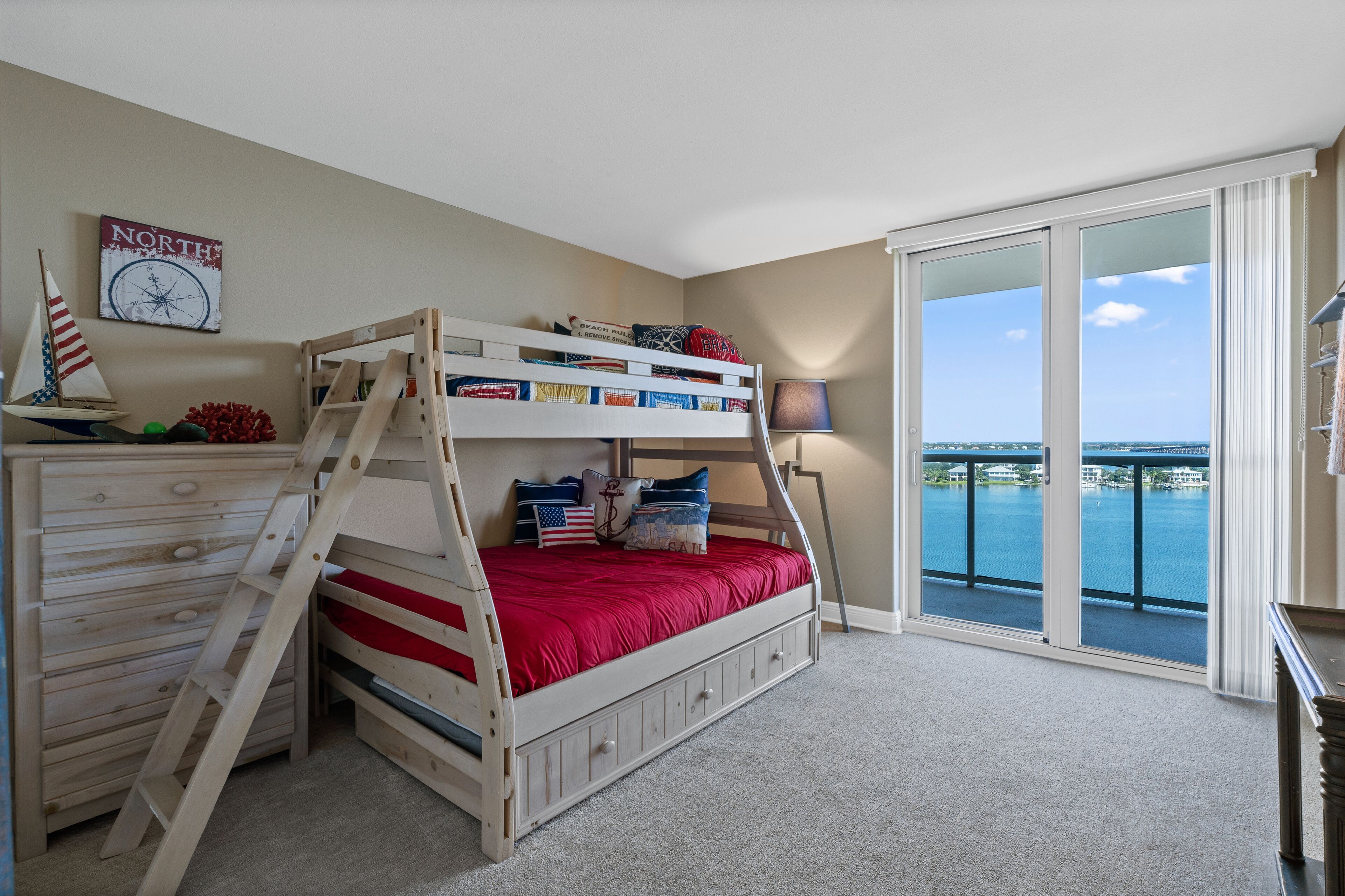 A bunk bed with a ladder, a dresser, a clock, and a view of a body of water through large windows.