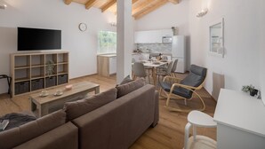 A modern living room with a sofa, coffee table, and TV. There is a kitchen area with a dining table and chairs. The room has wooden beams on the ceiling and large windows.