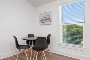 A dining area with a round table and four chairs, a window with blinds, and a painting on the wall.