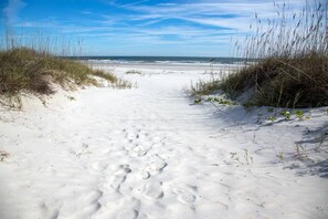 A sandy beach path leading to the ocean with footprints and sparse vegetation.