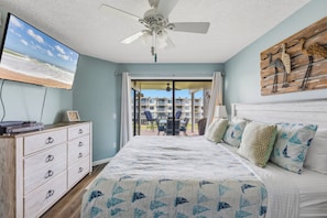A bedroom with a bed, a ceiling fan, a TV, a dresser, and a view of the outside.
