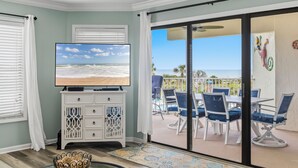 A room with a TV, a white cabinet, a patterned rug, and a balcony with chairs and a table. - Picture Perfect at Colony Reef Club 1310 (St. Augustine)