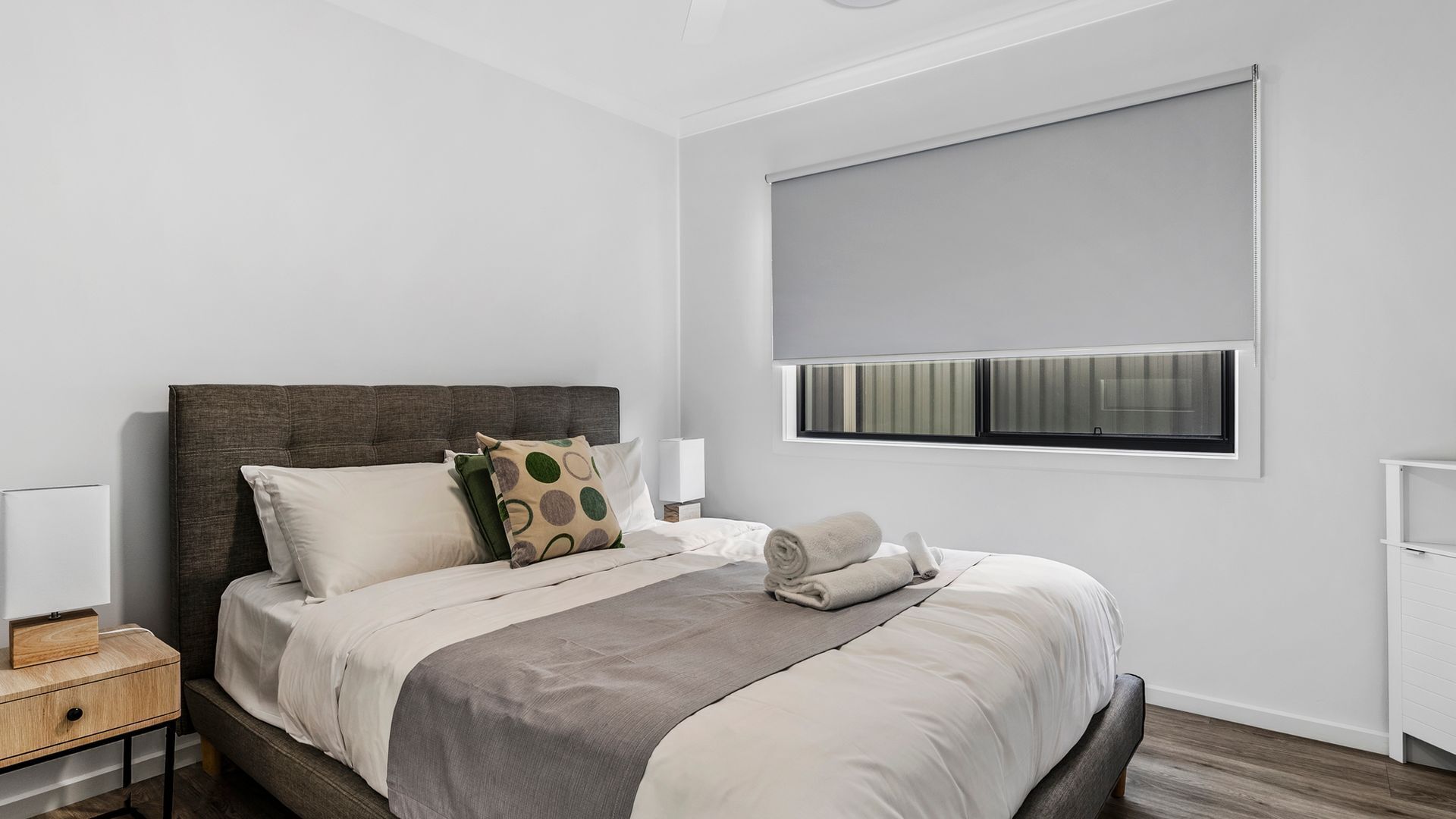 A modern bedroom with a large bed, a bedside table, a nightstand, a window with blinds, and a view of the outside.