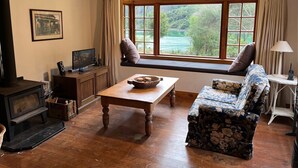 A cozy living room with a wooden floor, a fireplace, a television, a bench by the window, and a floral sofa. - Private seaside cottage with boat only access. (Havelock)