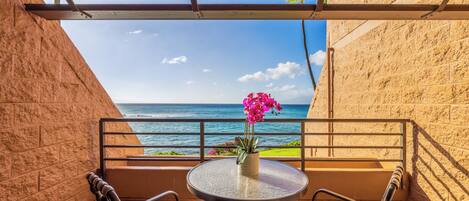 Un balcón con vistas al océano, una mesa redonda con sillas y un jarrón con flores rosadas.