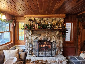 A cozy room with a stone fireplace, wooden ceiling, and a window with a view of trees.