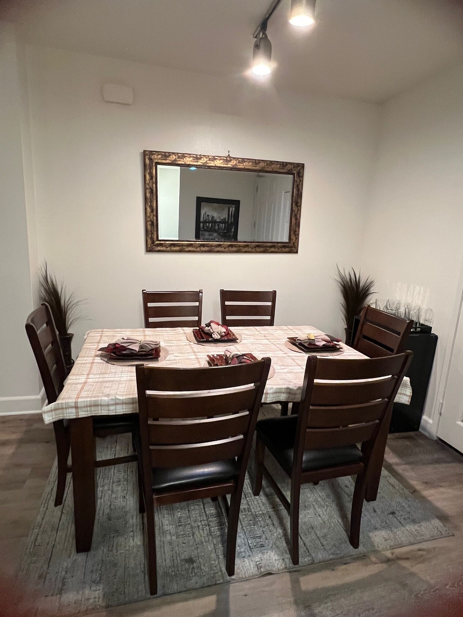 A dining room with a rectangular table, six wooden chairs, a mirror on the wall, and two potted plants.