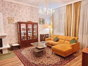 Uma sala de estar com sofá, mesa de centro, lareira, lustre e uma grande janela com cortinas.