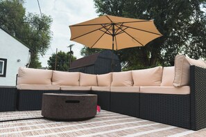 A sectional sofa with a large umbrella, a round ottoman, and a patterned tablecloth. - Traveler’s Dream Home! Hot tub, Firepit, & BBQ (Colorado Springs)