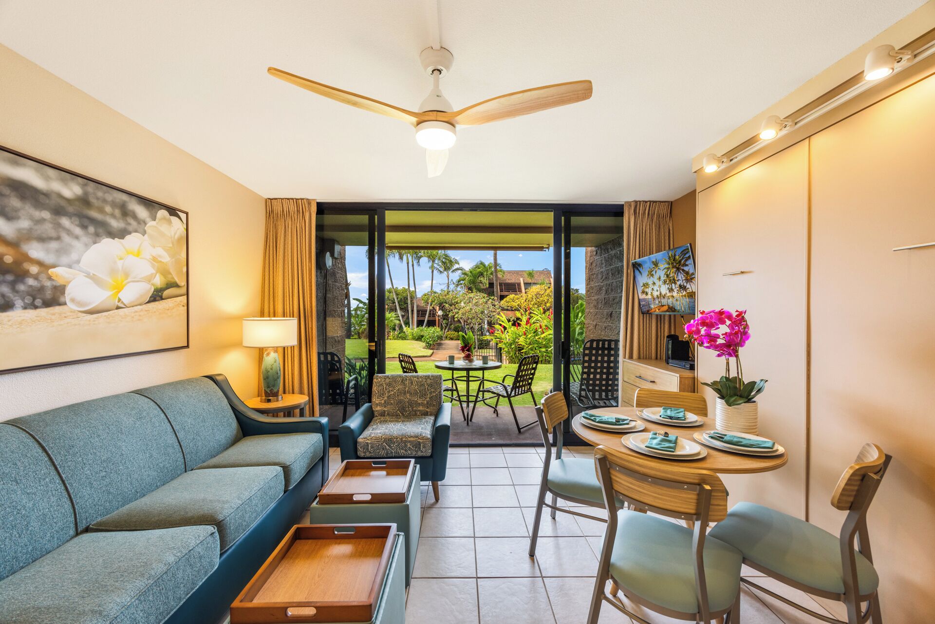 A hotel room with a sofa, a dining table with chairs, a ceiling fan, and a view of a garden with palm trees.