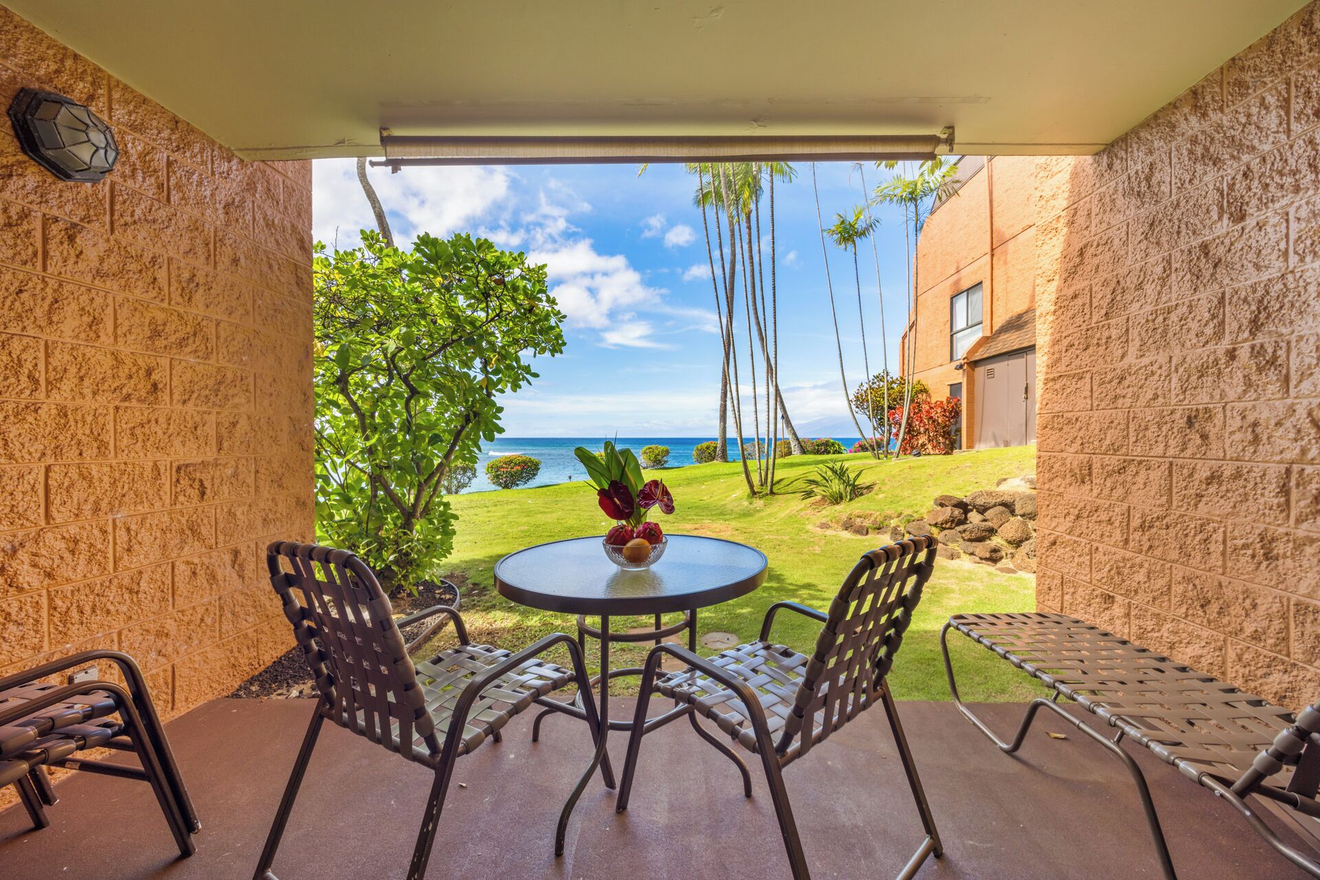 A covered patio with a round table and four chairs, overlooking a lawn with a view of the ocean and a building in the background.