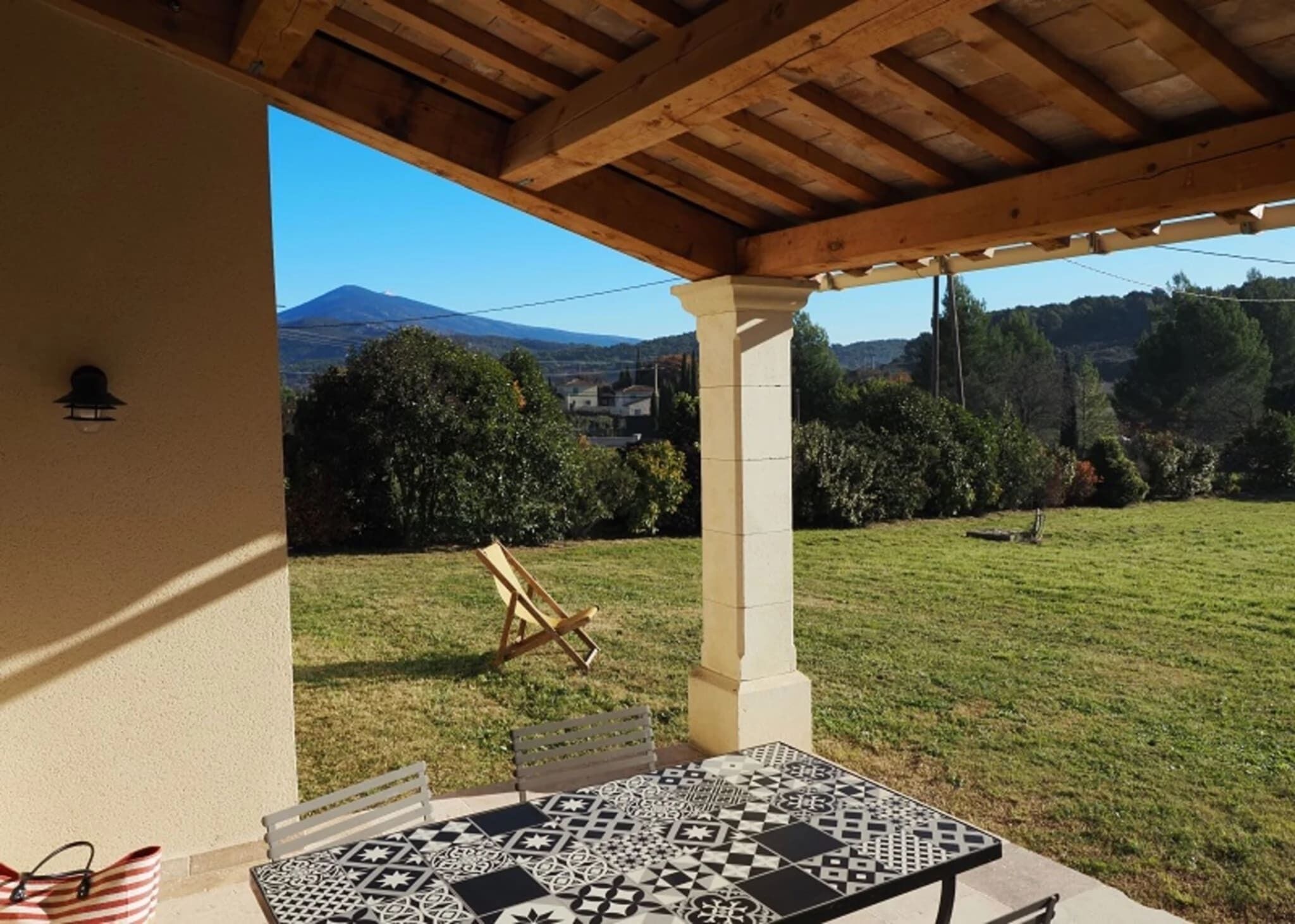 An outdoor patio with a table and chairs, a wooden pergola, and a view of a grassy area with trees and a mountain in the background.