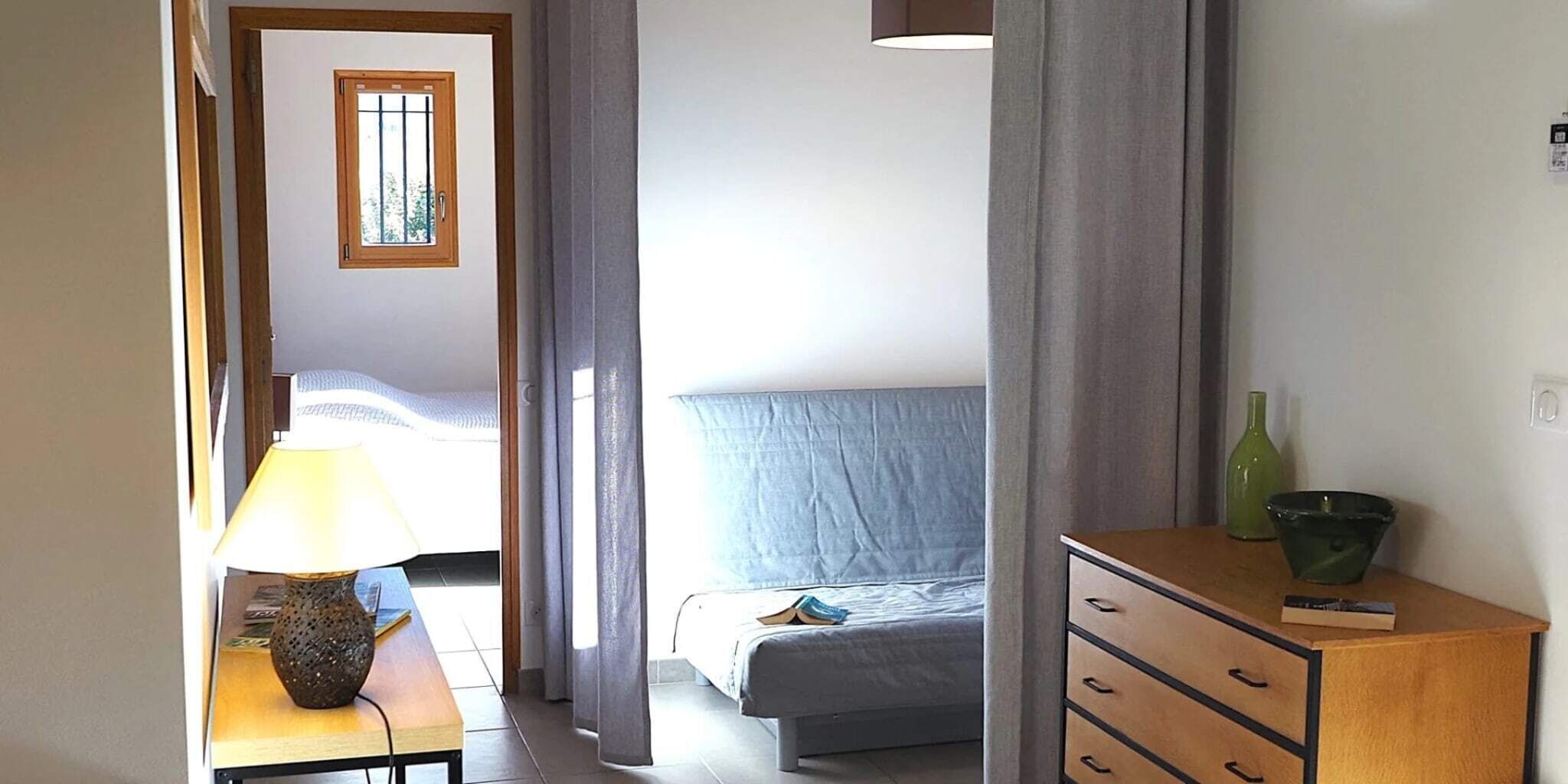 A bedroom with a bed, a bedside table with a lamp, a dresser, and a window with curtains.