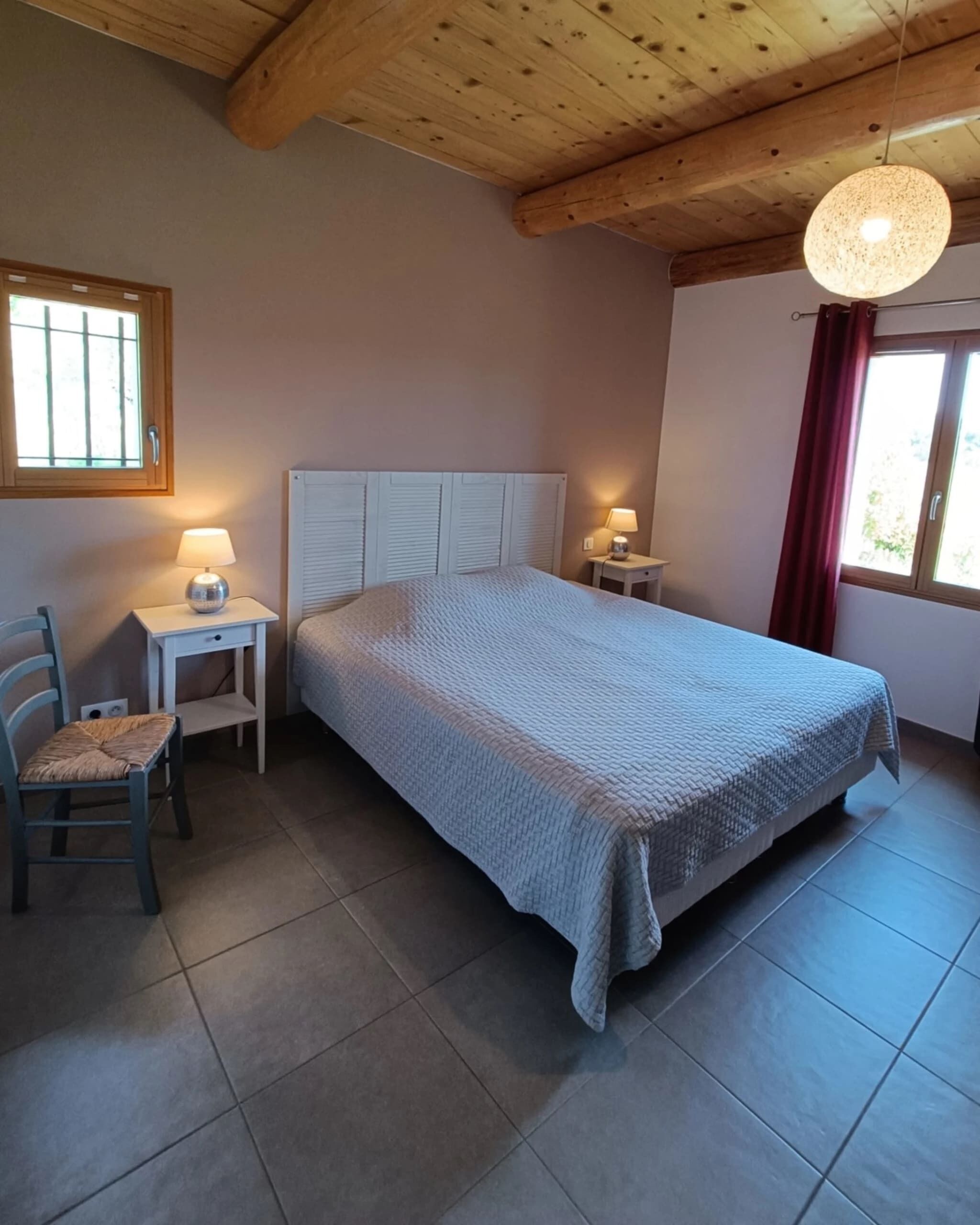 A bedroom with a bed, two bedside tables, a chair, a window with curtains, and a wooden ceiling.