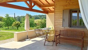 A wooden cabin with a patio area featuring a table and chairs, a bench, and a window overlooking a forested landscape. - Villa Garance with air conditioning (bedroom, living room, and kitchen) (LE BARROUX)