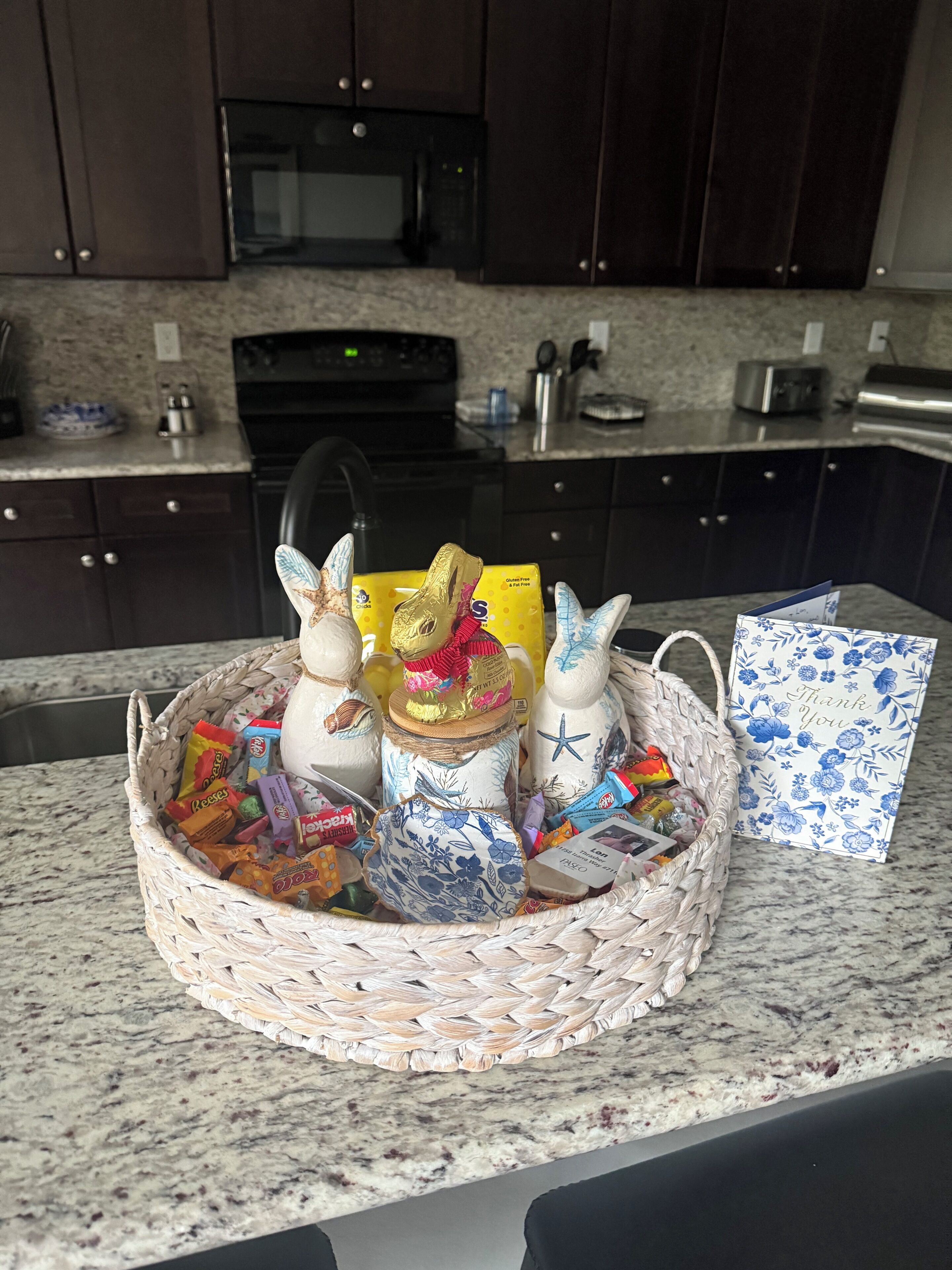A kitchen counter with a woven basket containing various items, including a jar with a lid, a decorative item resembling a rabbit, and a thank you card.