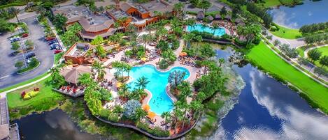 An aerial view of a resort with a central swimming pool, surrounded by lush greenery and palm trees.