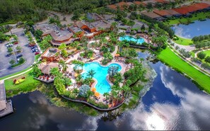 An aerial view of a resort with a central swimming pool, surrounded by lush greenery and palm trees.