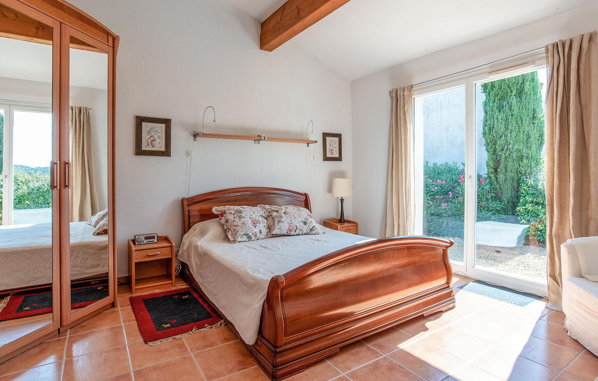 A bedroom with a wooden bed, a nightstand with a radio, a window with a view of greenery, and a sliding door leading to an outdoor area.