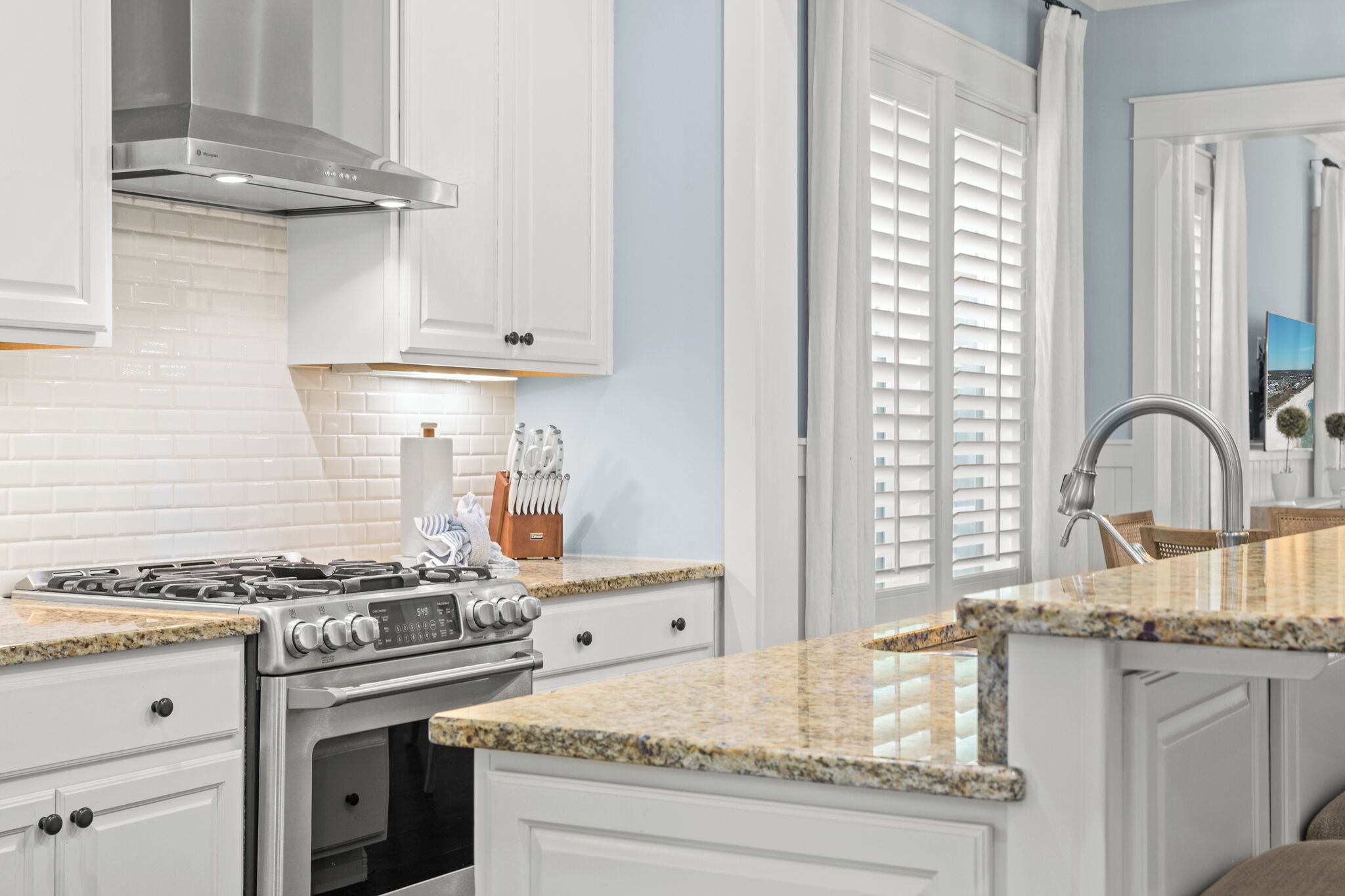 A kitchen with white cabinets, a stainless steel range hood, a granite countertop, and a window with blinds.