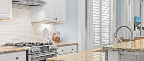 A kitchen with white cabinets, a stainless steel range hood, a granite countertop, and a window with blinds.