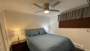 Ein Schlafzimmer mit Bett, Nachttisch, Lampe, Deckenventilator und Fenster mit Jalousien.