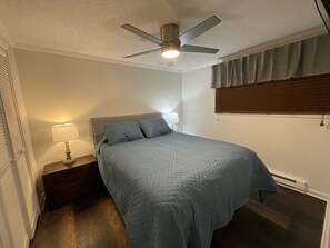 Une chambre à coucher comprenant un lit, une table de chevet, une lampe, un ventilateur de plafond et une fenêtre avec des stores.