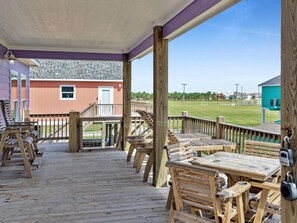A wooden deck with outdoor seating, a covered area, and a view of a building and grassy area. - Egret Escape-Sleeps 14, Park 1 House Away, Pet & Kid Friendly, Built 2022 (Bolivar Peninsula)