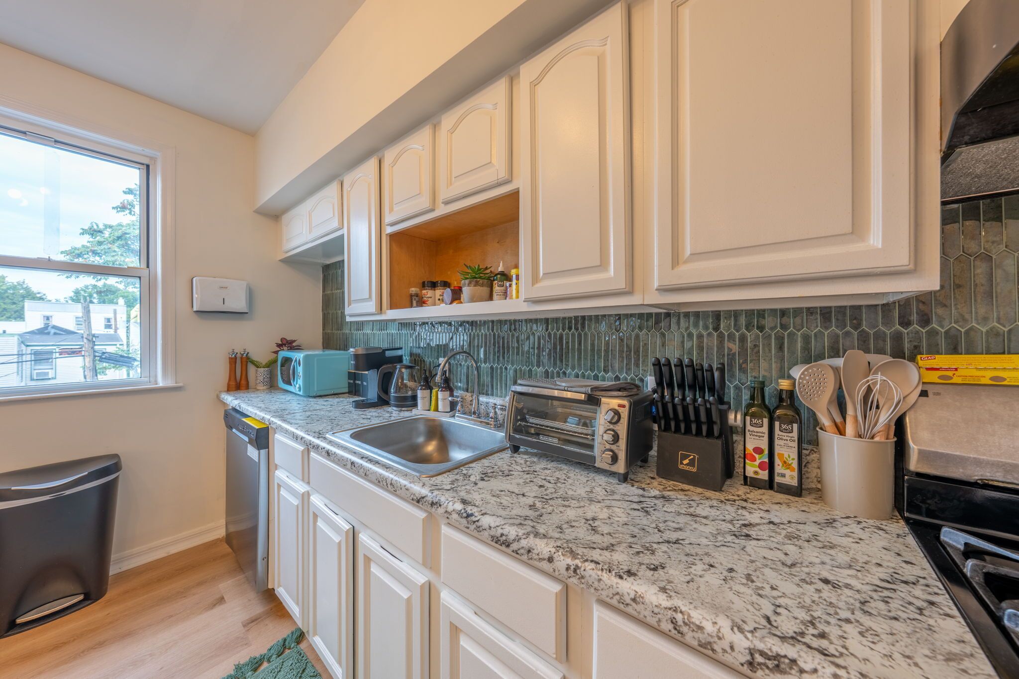 A kitchen with white cabinets, a granite countertop, a dishwasher, a sink, a toaster, and a window with a view of a house.