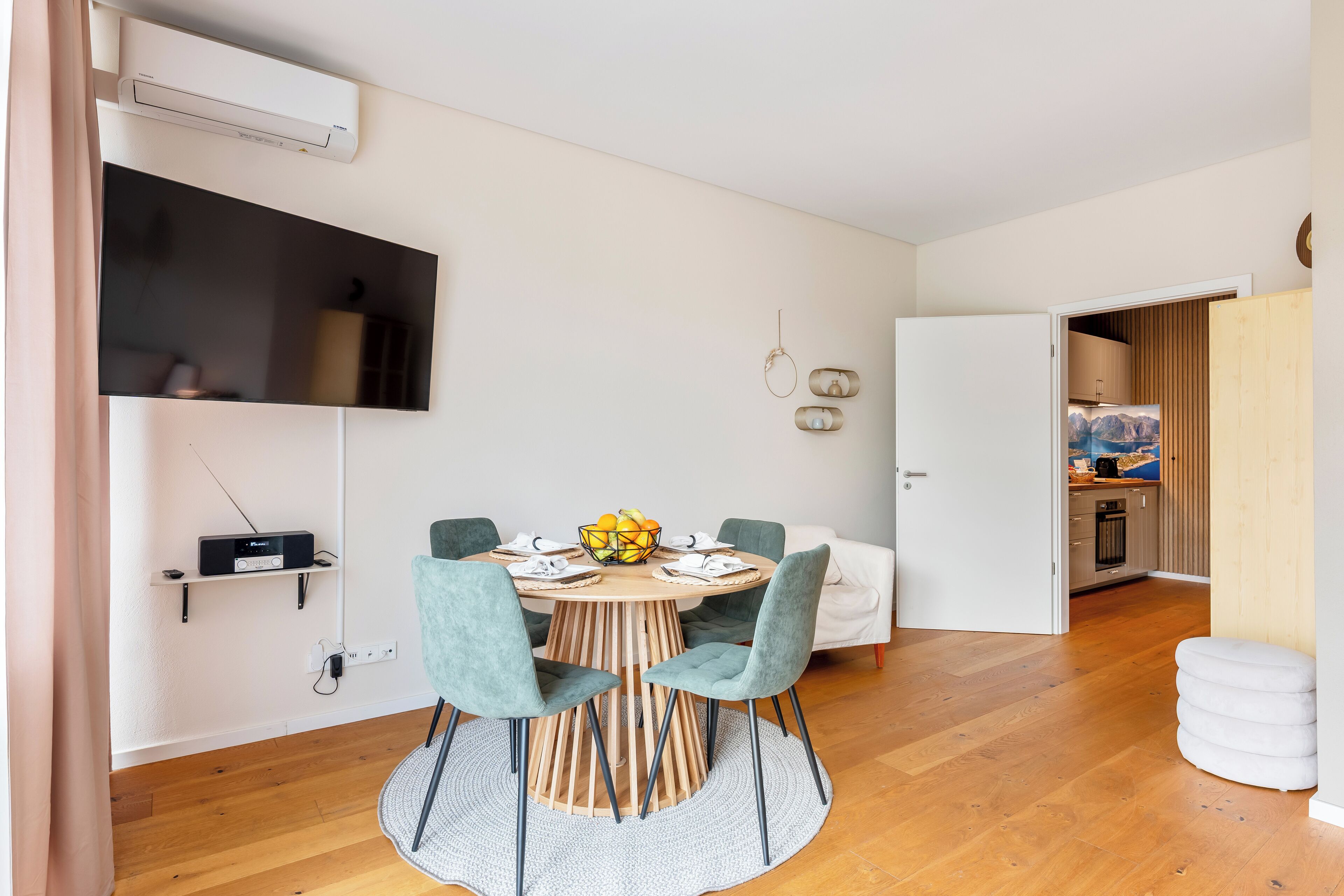 A modern dining area with a round table, four chairs, a TV mounted on the wall, and a kitchen visible through an open door.
