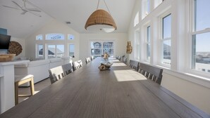 A spacious dining area with a long wooden table, white walls, large windows, and a ceiling fan. - 7138- Legasea, - Coastal Escape w/ Ocean Views & Private Pool (Waves)