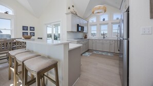 A modern kitchen with white cabinetry, a central island with bar stools, a built-in microwave, and a large refrigerator. - 7138- Legasea, - Coastal Escape w/ Ocean Views & Private Pool (Waves)