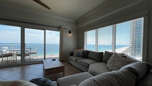 Un vaste espace de vie comprenant un grand canapé gris, une petite table et un balcon avec vue sur l’océan.