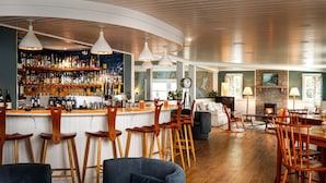 A bar area with wooden stools, a bar counter with a variety of bottles, and a seating area with a fireplace. - The Crocker House Country Inn (Rm 3) (Hancock)
