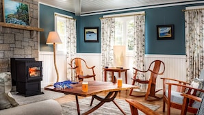 A cozy living room with a stone fireplace, wooden furniture, and a view of the outdoors through large windows. - The Crocker House Country Inn (Rm 3) (Hancock)