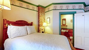 A neatly made bed with white linens, a wooden headboard, and a nightstand with a lamp. A brick wall and a door leading to a bathroom are visible. - The Crocker House Country Inn (Rm 6) (Hancock)
