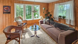 A cozy living room with a wicker chair, a sofa, and a glass coffee table. There are large windows with curtains, a lamp, and a framed picture on the wall.
