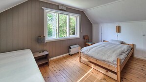 A bedroom with a sloped ceiling, a wooden bed, a chair, a radiator, and a window with a view of trees.