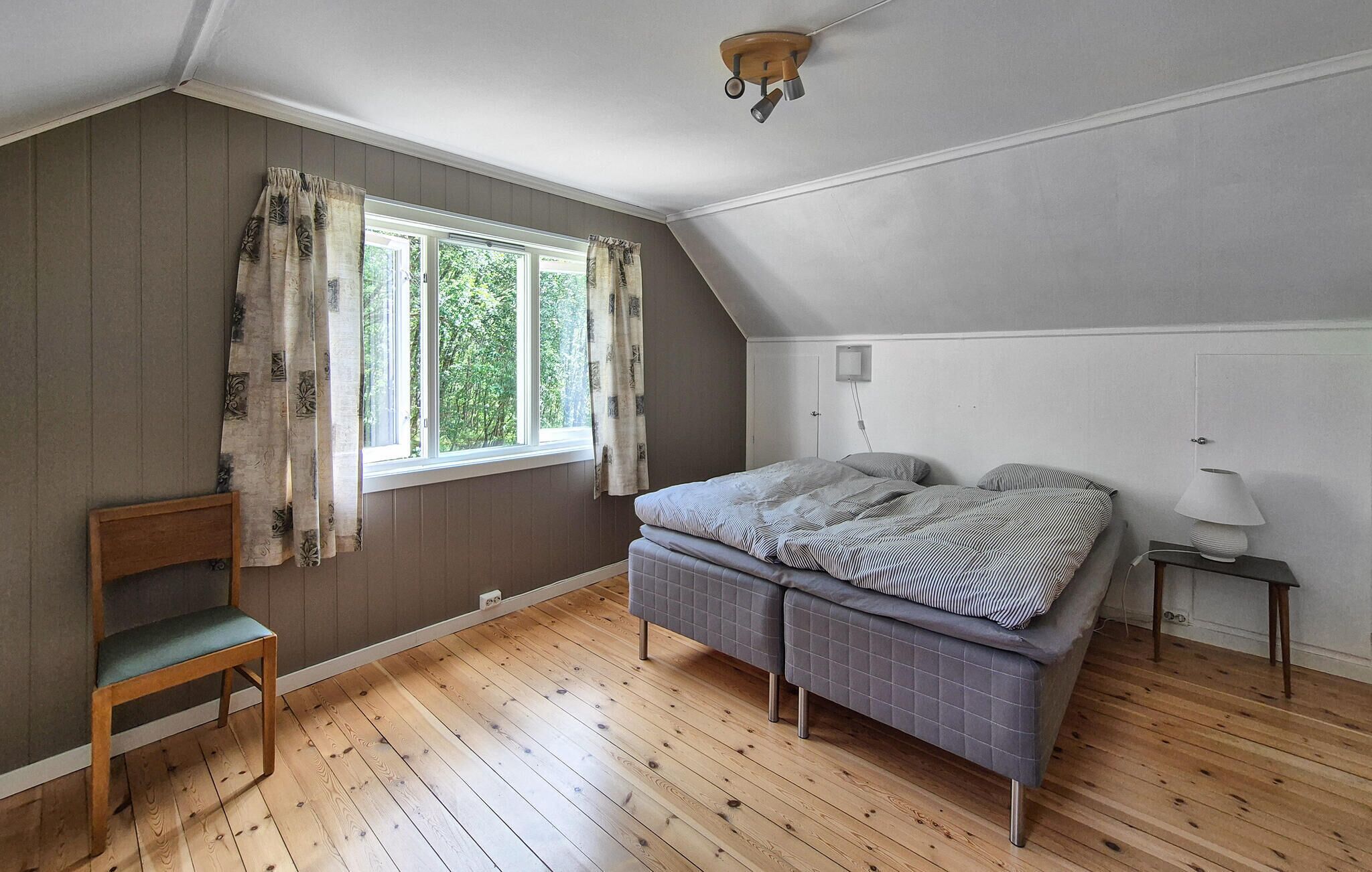 A bedroom with a bed, a chair, a bedside table, a lamp, and a window with curtains.