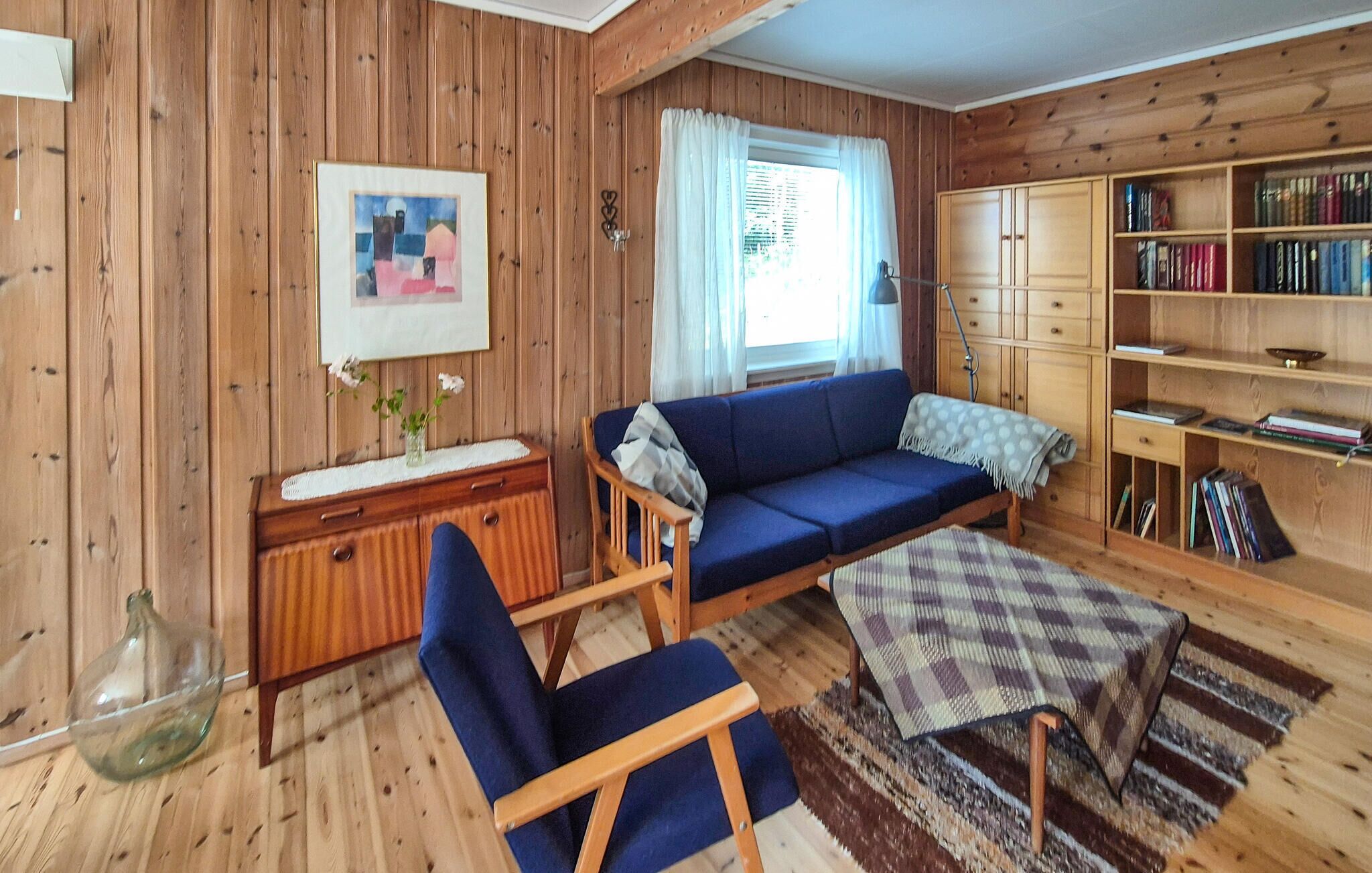 A cozy living room with wooden walls, a blue sofa, a wooden cabinet with books, and a small table with a checkered tablecloth.