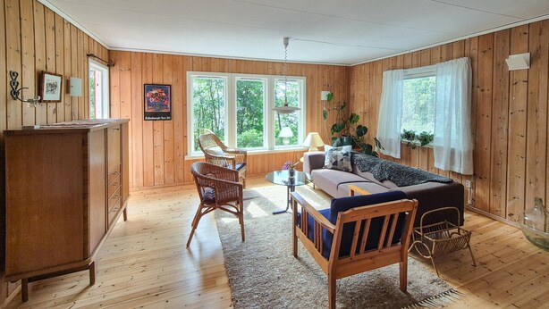 A cozy living room with a sofa, chairs, a small table, and a wooden cabinet. There are windows with curtains, a ceiling fan, and a framed picture on the wall.