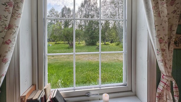 Una ventana con vista a un campo cubierto de hierba y árboles, una vela en el alféizar y libros apilados junto a la ventana.