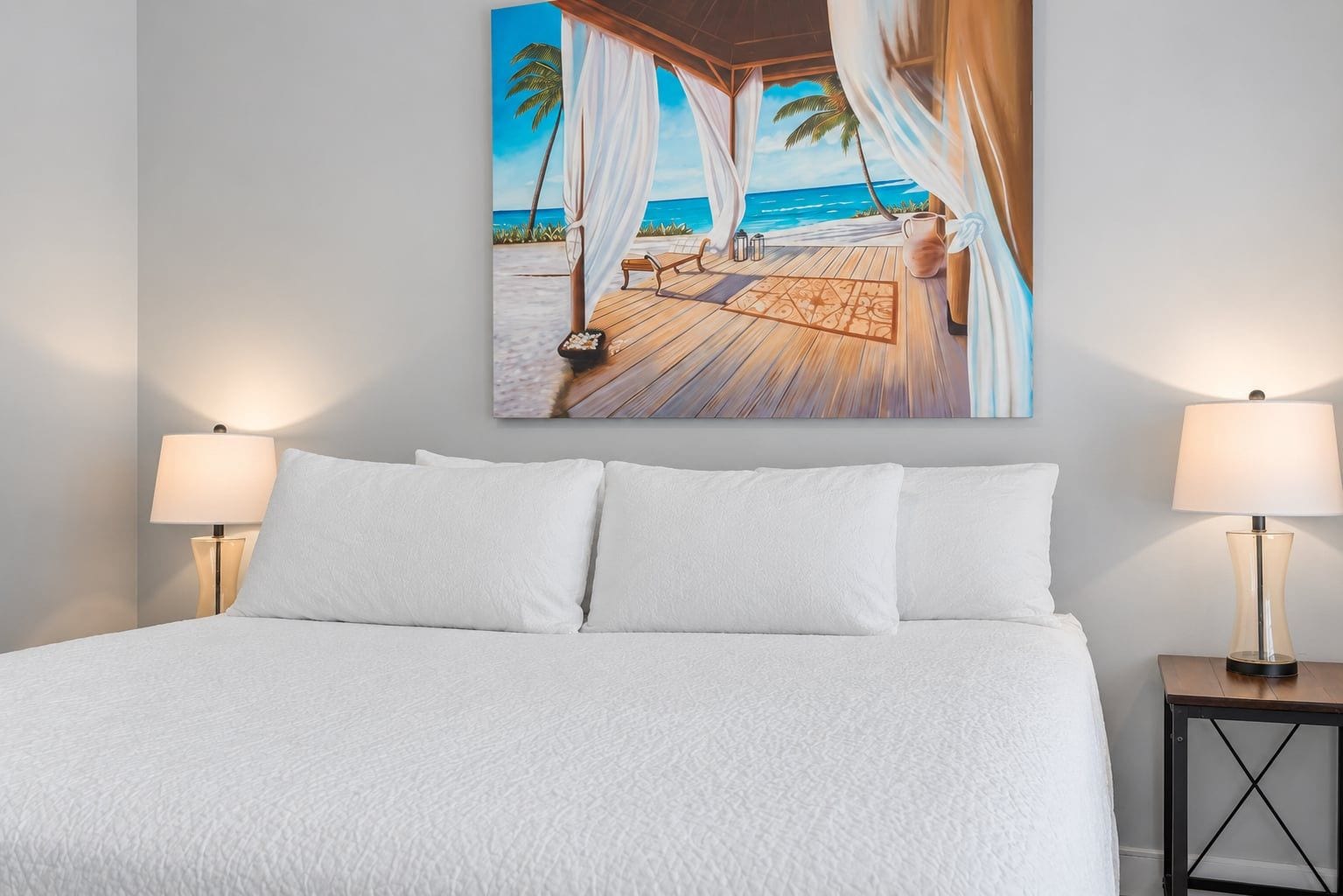 A bed with white bedding, two bedside lamps, and a painting of a beach scene.