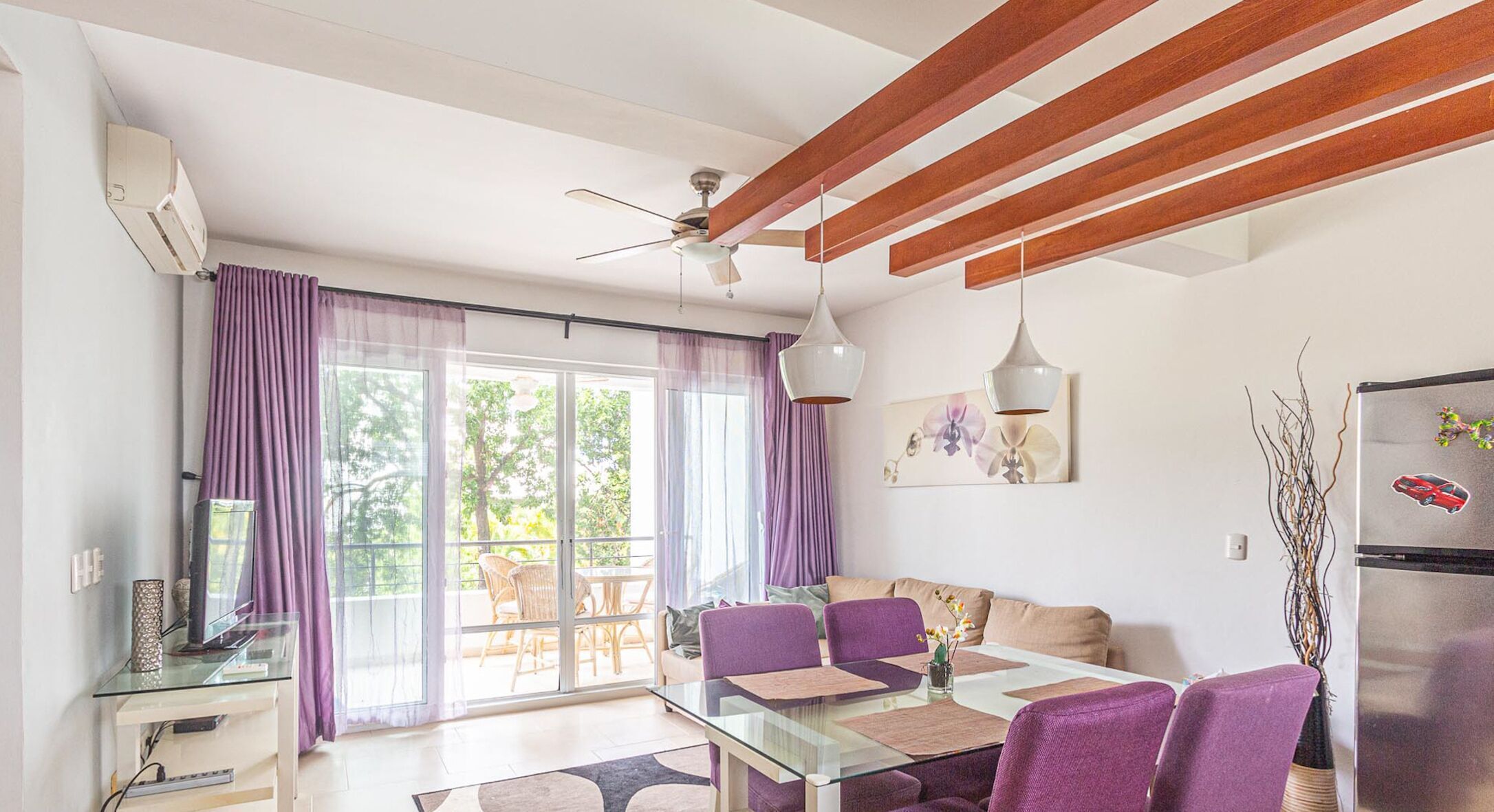 A modern living room with a glass dining table, purple chairs, a ceiling fan, and a large window with purple curtains.