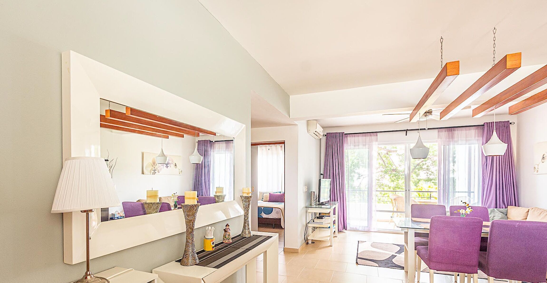 A modern living room with a large mirror, a dining area with purple chairs, and a TV.