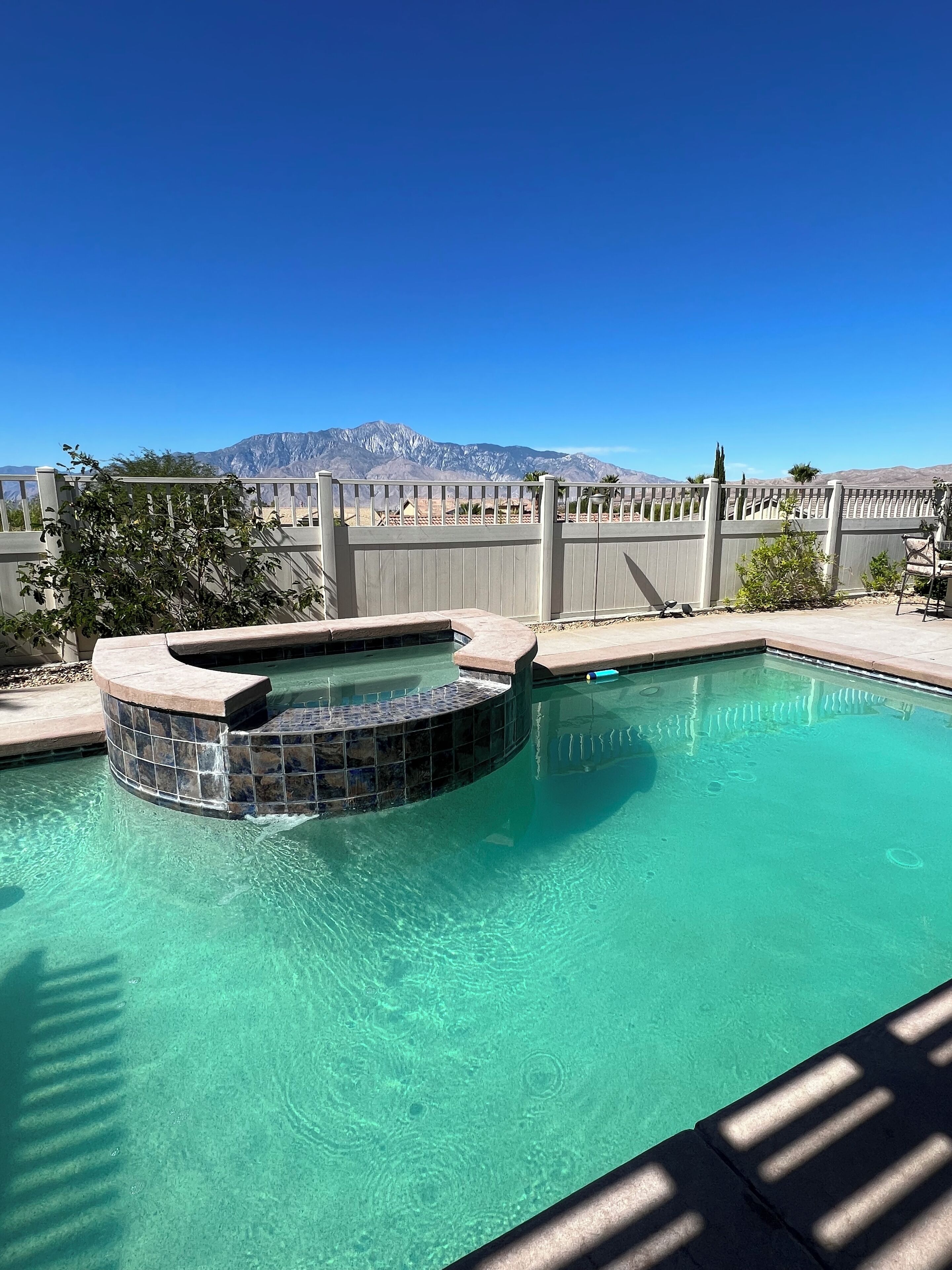 A swimming pool with a hot tub, surrounded by a wooden fence and a view of mountains.