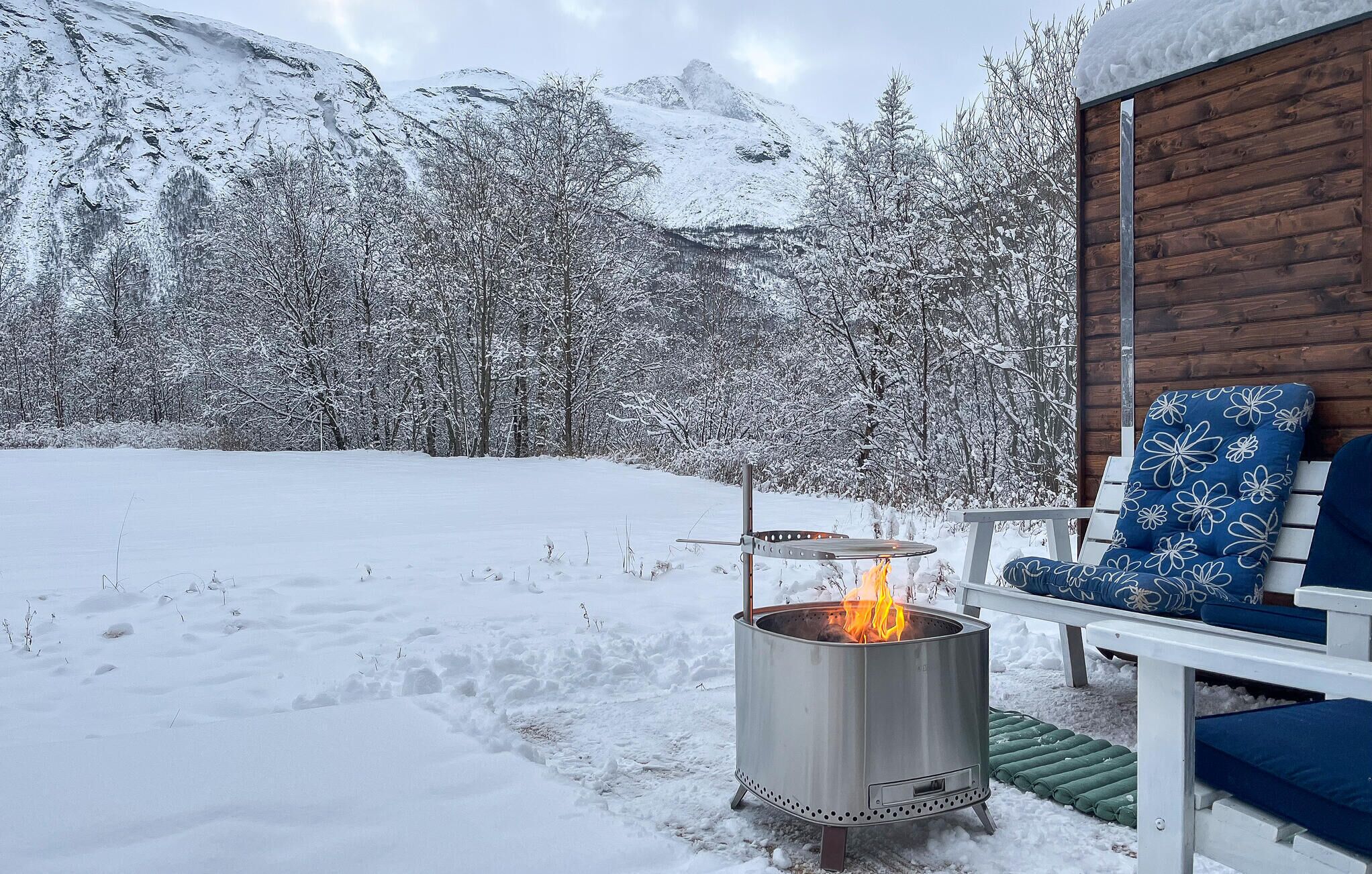 A metal fire pit with flames, a wooden cabin with a blue chair, and a snowy mountain backdrop.