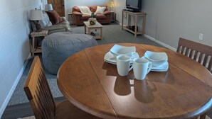 Un salón acogedor con una mesa redonda de madera dispuesta para comer, un sofá, un televisor y una puerta que da a otra habitación.